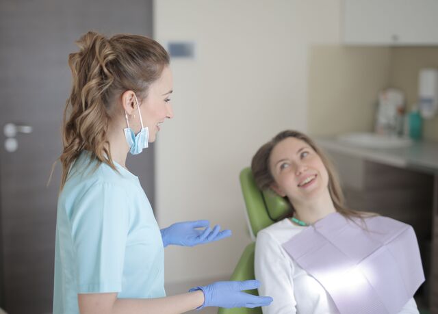 a dental professional in scrubs and gloves talks to a patient sitting in a dental chair who is smiling and wearing a protective bib